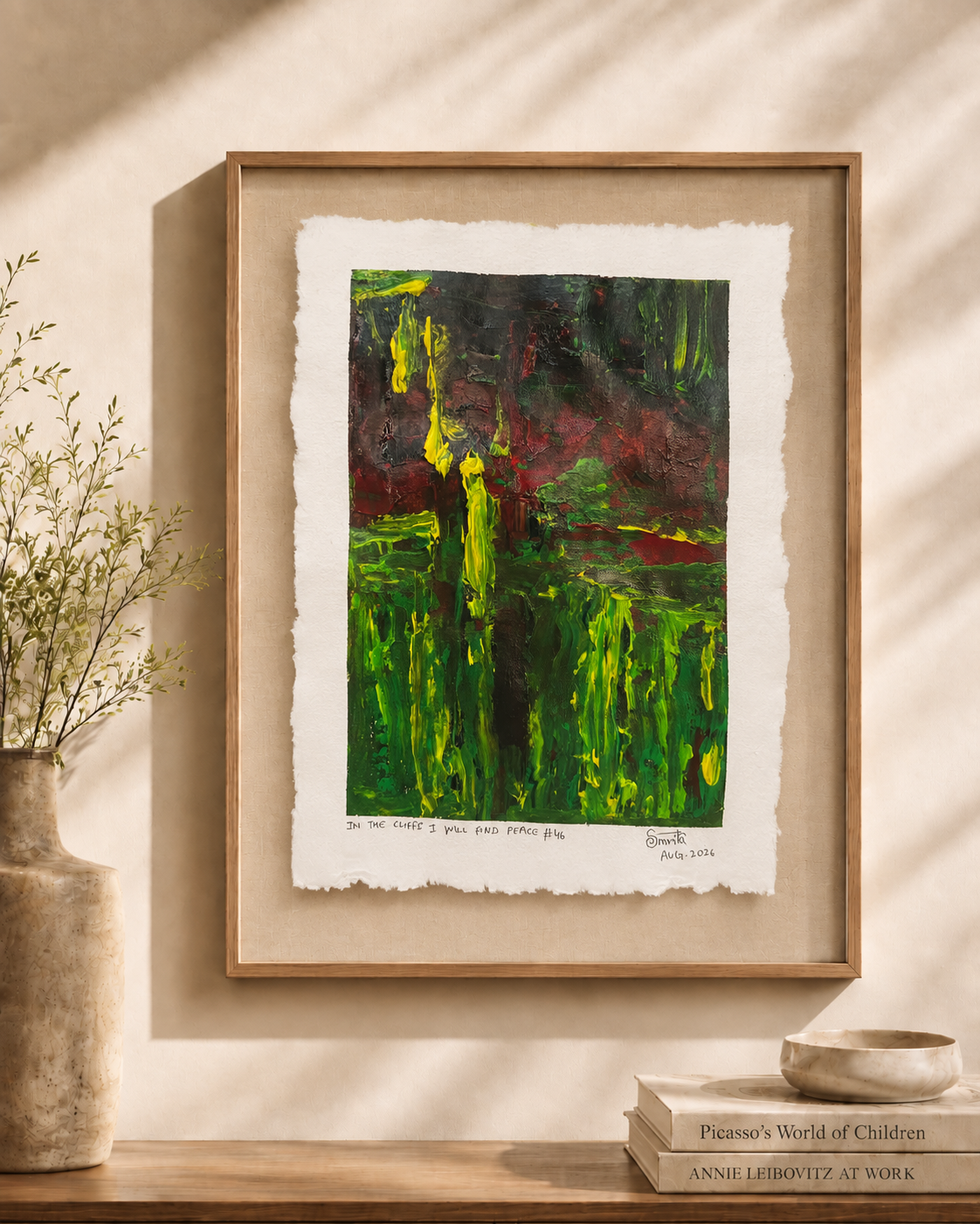 A framed abstract art print featuring vibrant green, yellow, and dark red brushstrokes on textured white paper, displayed on a wooden shelf next to a vase and books.
