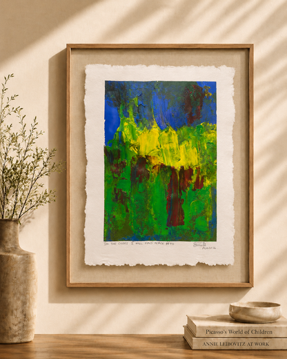 A framed abstract art print featuring vibrant green, yellow, and blue brushstrokes, styled on a wooden shelf next to a vase with dried branches and a stack of books under warm, sunlit shadows.