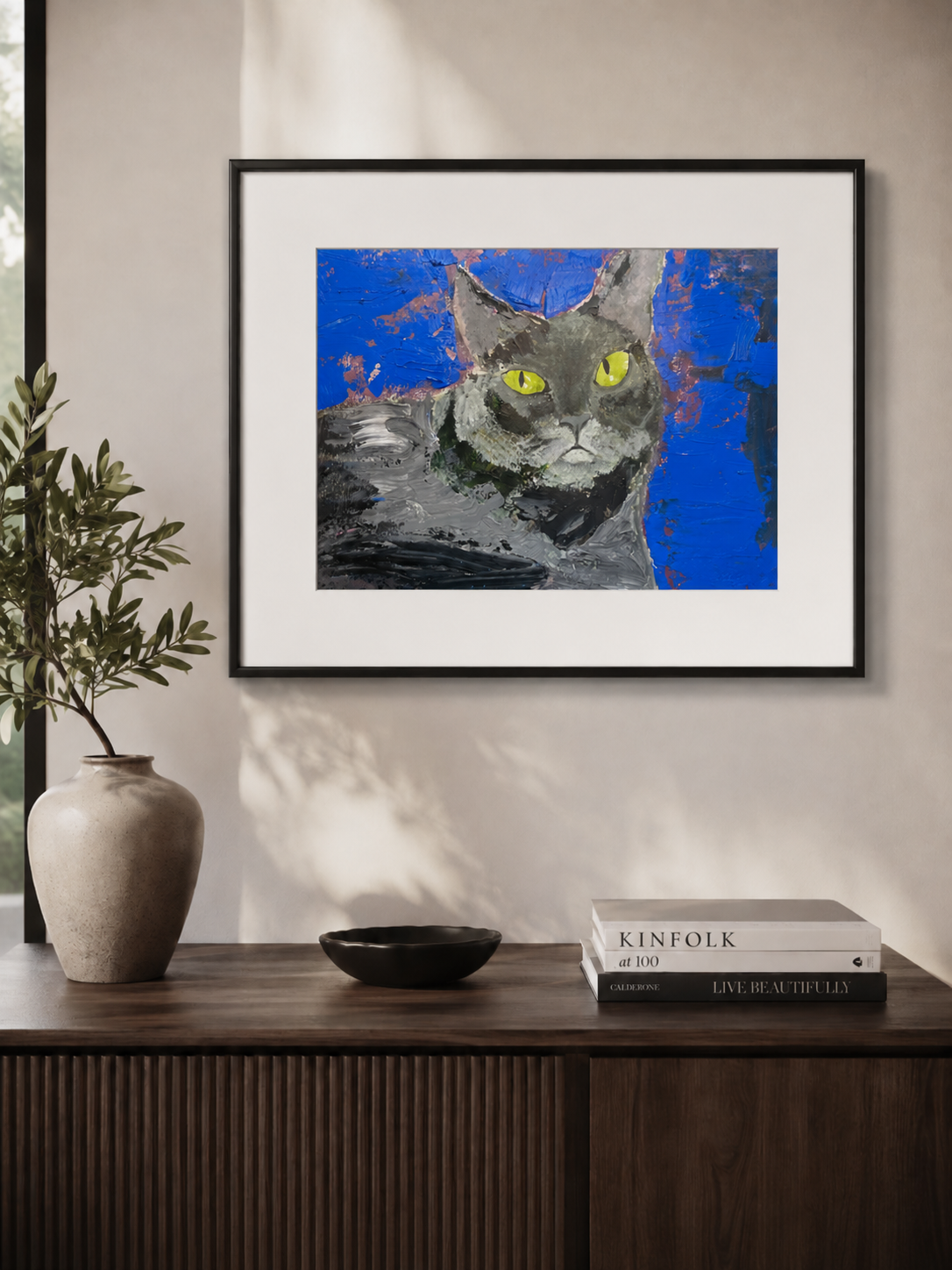 A framed art print of a gray cat with bright yellow eyes against a textured blue background, displayed on a beige wall above a dark wood console table with a vase and books.