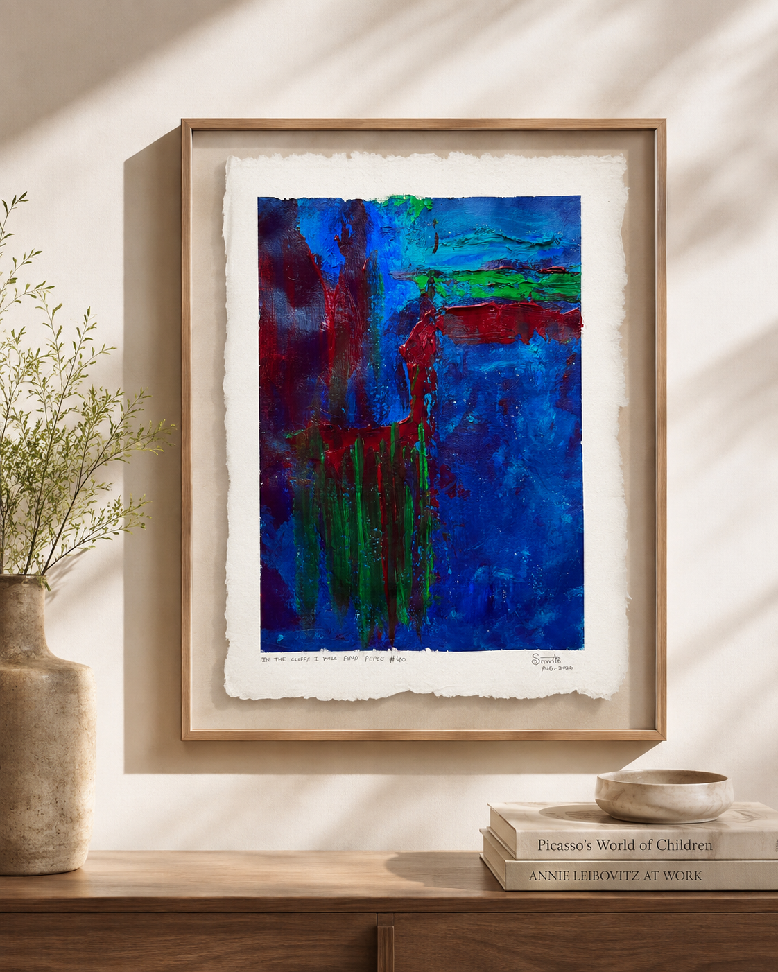 A framed abstract art print featuring vibrant blue, red, and green brushstrokes on textured white paper, displayed on a wooden console table in a styled room setting with a vase and books.