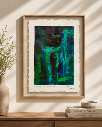 A framed abstract art print featuring vibrant green, teal, and dark blue brushstrokes on textured off-white paper, displayed in a light wood frame on a beige wall above a wooden console table with a vase and books.