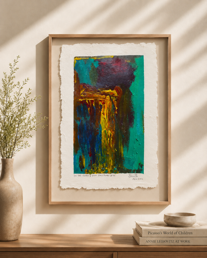 A framed abstract art print with textured, torn paper edges hangs on a beige wall above a wooden console table. The print features a vibrant, multicolored composition with deep teal, gold, yellow, and dark purple tones.
