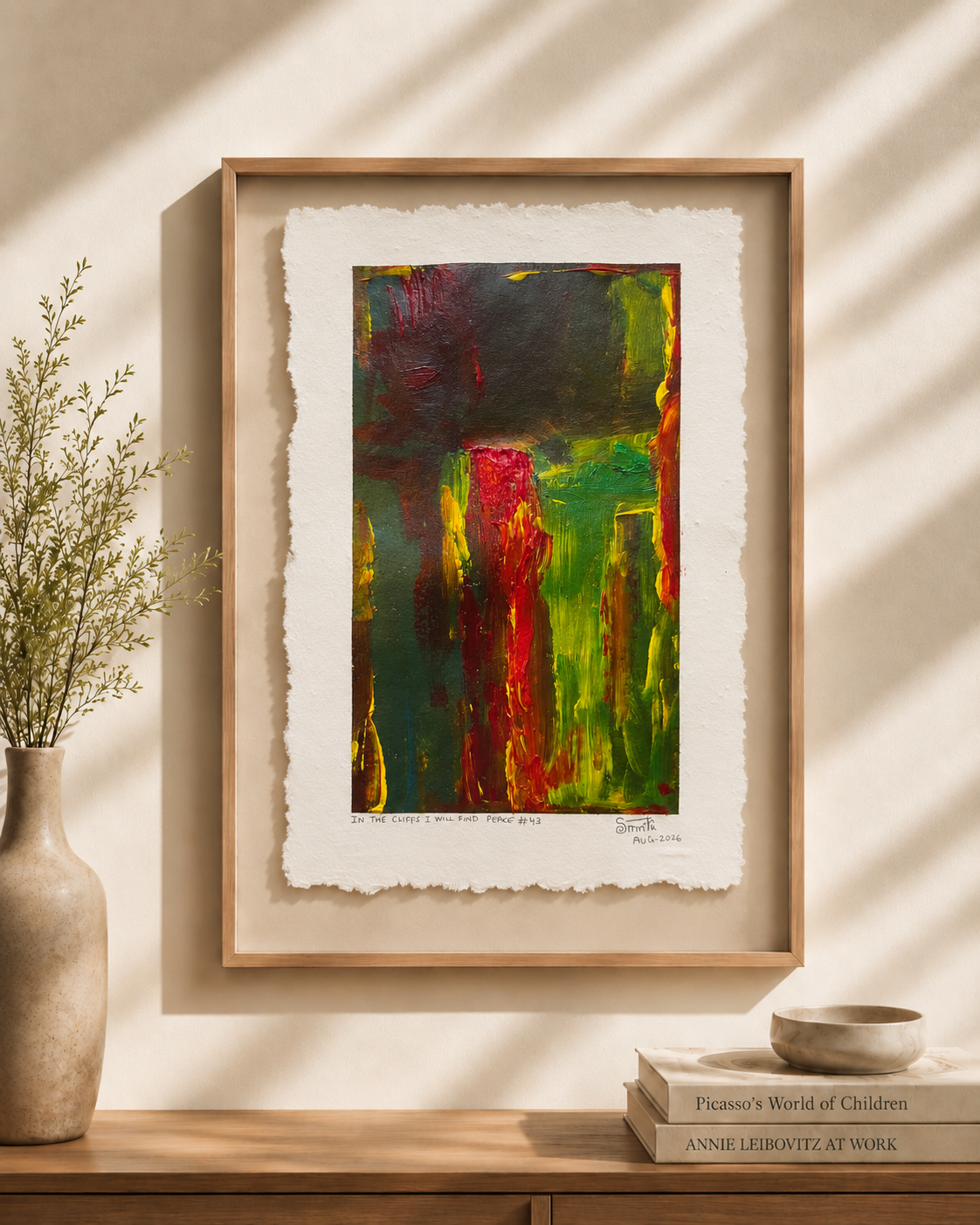 Framed abstract art print with textured white paper margins, featuring bold green, red, yellow, and dark green brushstrokes, displayed on a wooden console against a sunlit beige wall with a vase and books nearby.