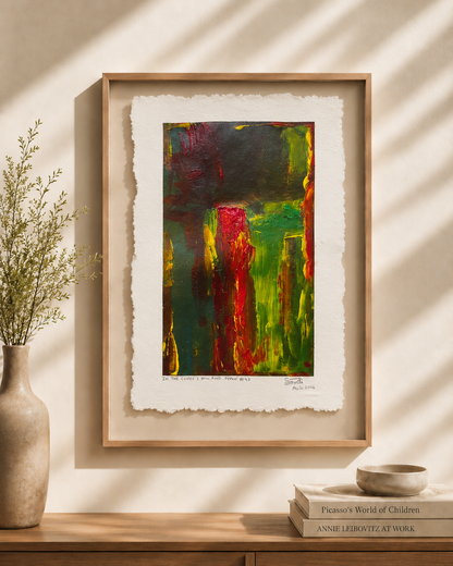 Framed abstract art print with textured white paper margins, featuring bold green, red, yellow, and dark green brushstrokes, displayed on a wooden console against a sunlit beige wall with a vase and books nearby.