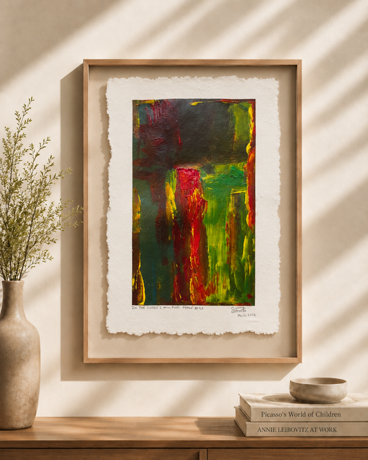 Framed abstract art print with textured white paper margins, featuring bold green, red, yellow, and dark green brushstrokes, displayed on a wooden console against a sunlit beige wall with a vase and books nearby.