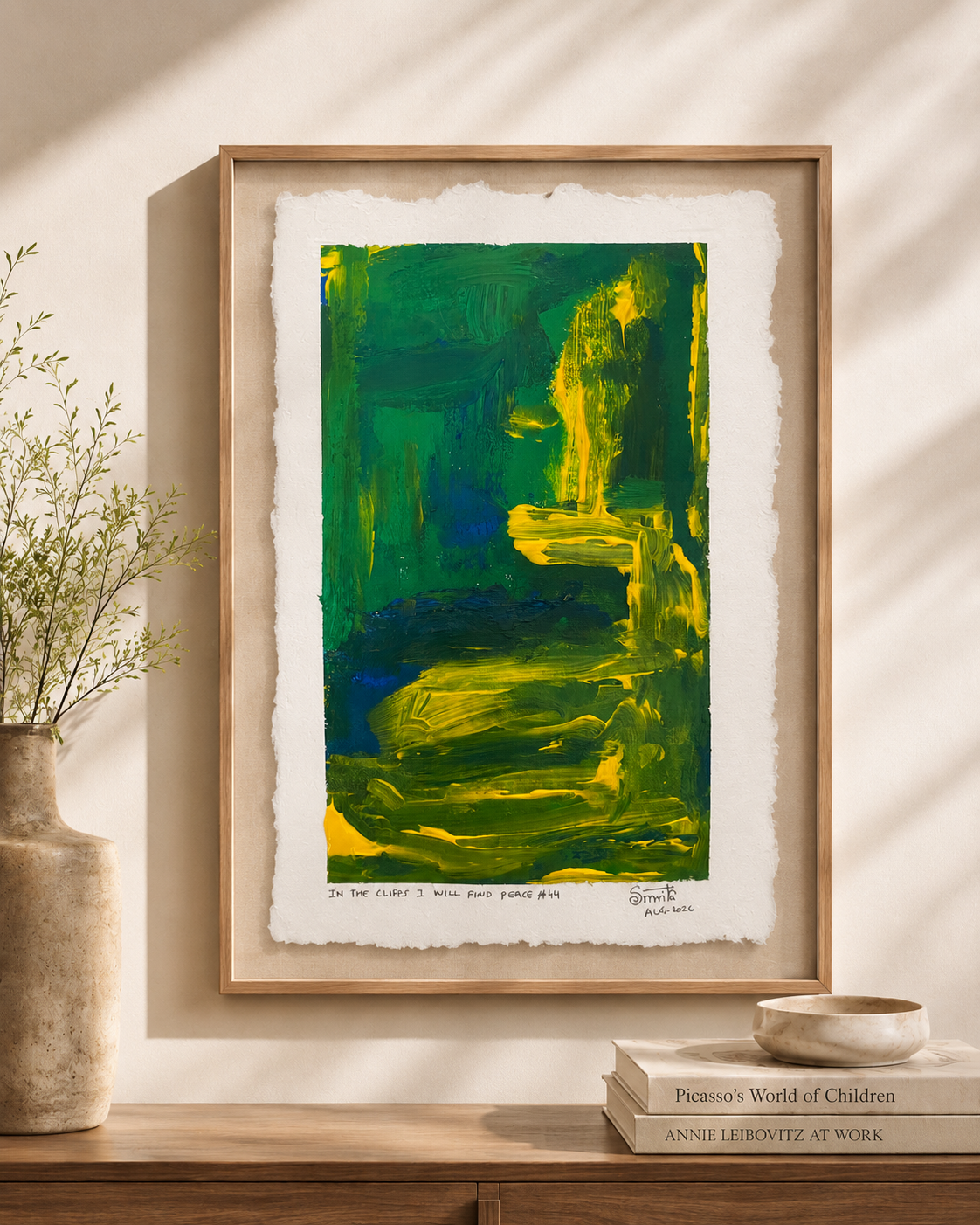 A framed abstract art print featuring bold green, yellow, and blue brushstrokes, displayed on a wooden console table with a vase and books in a sunlit room.