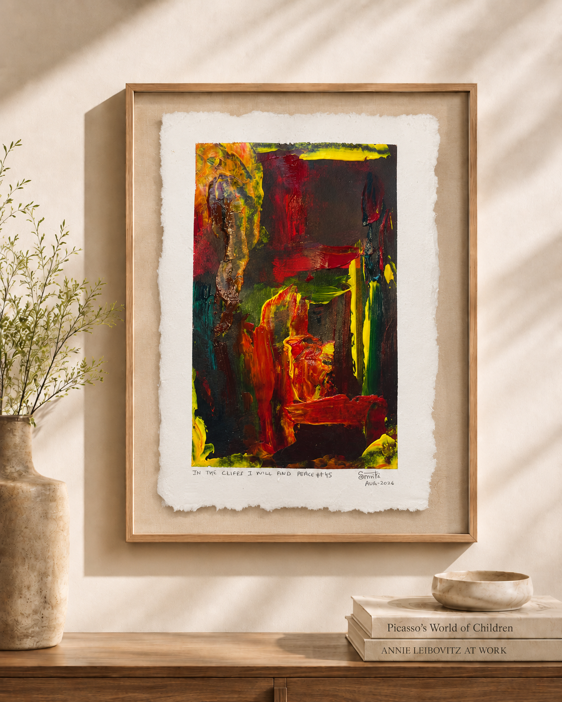 Framed abstract art print with a light wood frame, displayed on a beige wall above a wooden console table with a vase and books. The artwork features bold, expressive brushstrokes in red, green, yellow, and black on textured off-white paper.