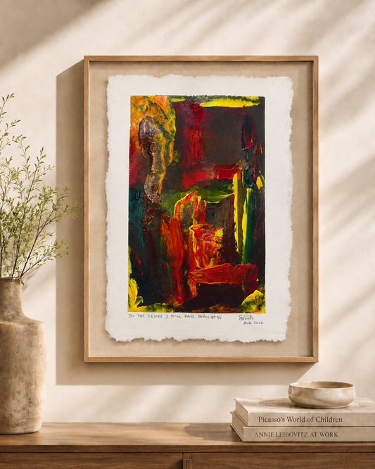 Framed abstract art print with a light wood frame, displayed on a beige wall above a wooden console table with a vase and books. The artwork features bold, expressive brushstrokes in red, green, yellow, and black on textured off-white paper.
