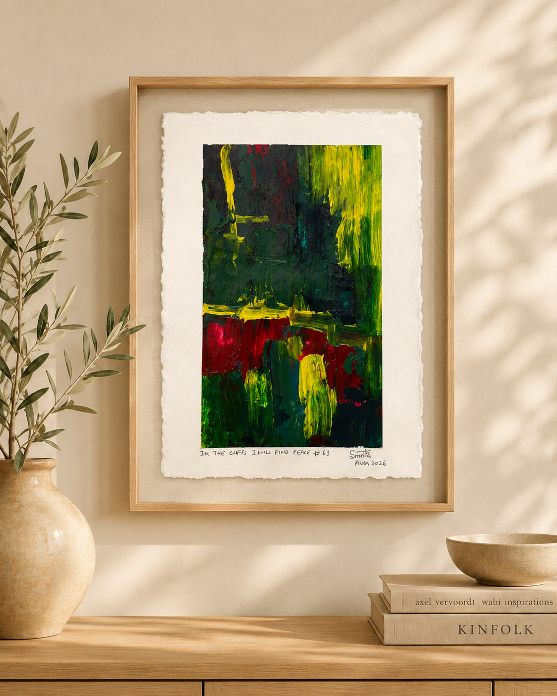 Framed abstract art print with a light wood frame, displayed on a wooden console table against a beige wall with soft leaf shadows. The artwork features bold green, yellow, red, and black brushstrokes on white paper with visible deckled edges.