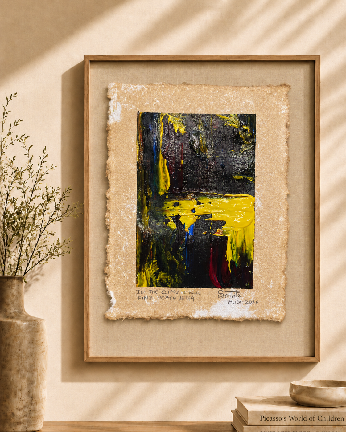 A framed abstract art print featuring bold black, yellow, and blue paint strokes on textured beige paper, displayed in a light wood frame on a warm beige wall with decorative shadows, a vase, and books.