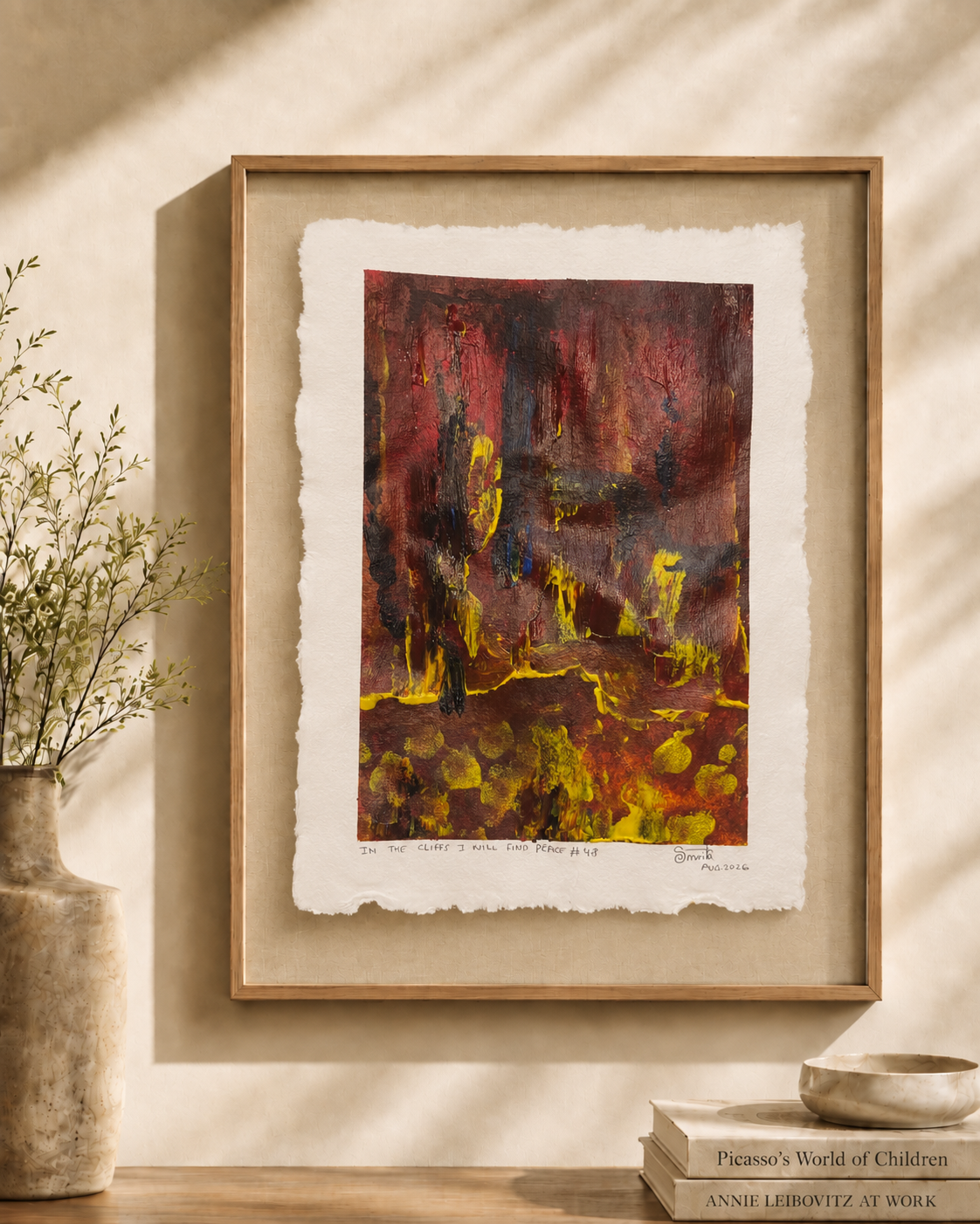 A framed abstract art print with a textured white border hangs on a beige wall in a styled interior setting with a vase of branches and books on a wooden surface.